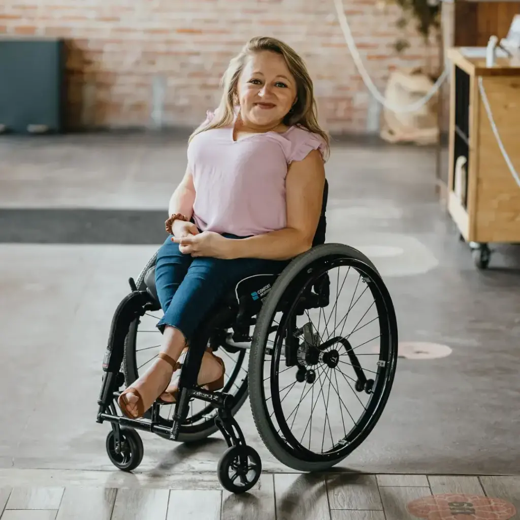 Emily Voorde. She has light skin, blonde hair, wears a pink shirt, jeans and brown sandals, and uses a wheelchair. She smiles. 