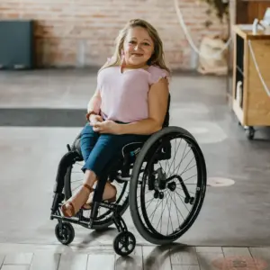 Emily Voorde. She has light skin, blonde hair, wears a pink shirt, jeans and brown sandals, and uses a wheelchair. She smiles.