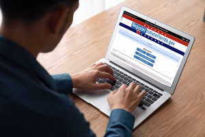 A person sits at a wooden desk using a laptop, registering to vote online on the RegisterToVoteFlorida.gov website. The screen displays options in both English and Spanish for new registrations and updates. The user’s hands are visible typing on the keyboard.